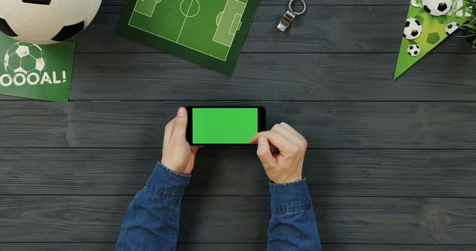 Top view on the black horizontal smartphone on the dark grey wooden desk of football trainer and male Caucasian hands scrolling and taping on the chroma key screen. Green screen.