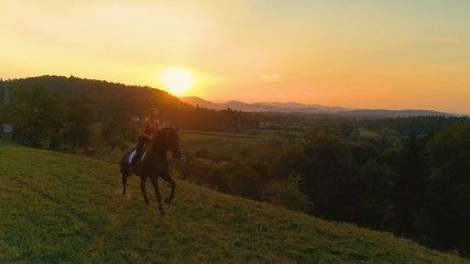 Fototapeta na wymiar LENS FLARE: Happy young woman galloping on her beautiful horse at idyllic sunset