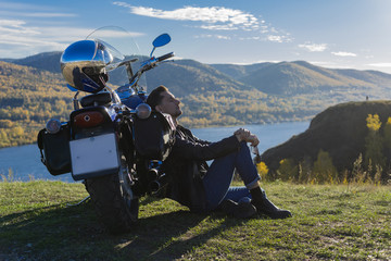Fototapeta na wymiar Young man wearing a black leather jacket and jeans sits outdoor on a ground at a motorcycle, resting on a mountain above the river. Lifestyle, travel. Copy space