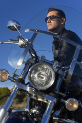 Young man wearing a black leather jacket, sunglasses and jeans sits outdoor on a motorcycle, on a mountain on a blue sky background. Lifestyle, travel. Copy space