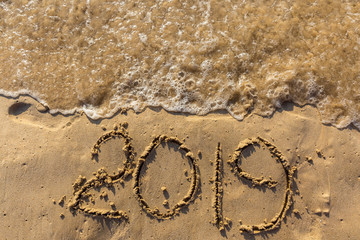 Concept of Happy New Year 2019 is coming and leaving year of 2018. Text on the sea beach and footprints on the sand. Wave water covering digits inscription. Meet at a tropical resort in Thailand.