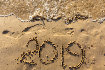 Concept of Happy New Year 2019 is coming and leaving year of 2018. Text on the sea beach and footprints on the sand. Wave water covering digits inscription. Meet at a tropical resort in Thailand.