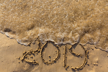 Concept of Happy New Year 2019 is coming and leaving year of 2018. Text on the sea beach and footprints on the sand. Wave water covering digits inscription. Meet at a tropical resort in Thailand.