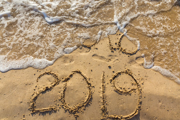 Concept of Happy New Year 2019 is coming and leaving year of 2018. Text on the sea beach and footprints on the sand. Wave water covering digits inscription. Meet at a tropical resort in Thailand.