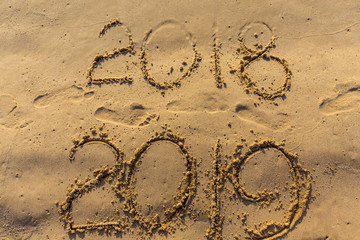 Concept of Happy New Year 2019 is coming and leaving year of 2018. Text on the sea beach and footprints on the sand. Wave water covering digits inscription. Meet at a tropical resort in Thailand.