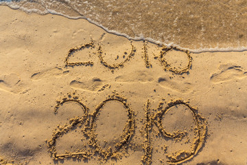 Concept of Happy New Year 2019 is coming and leaving year of 2018. Text on the sea beach and footprints on the sand. Wave water covering digits inscription. Meet at a tropical resort in Thailand.