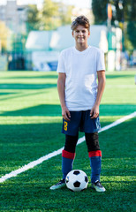 Obraz premium cute young boy in white blue sportswear stands next to classical black and white football ball on the stadium field. Soccer game, training, hobby concept. 