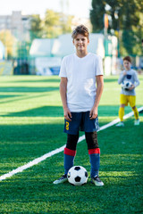 cute young boy in white blue sportswear stands next to classical black and white football ball on the stadium field. Soccer game, training, hobby concept. 