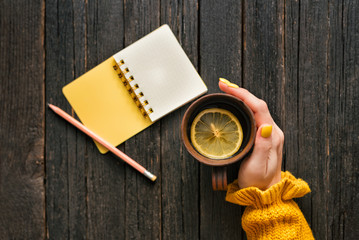 Mug of tea with lemon in a female hand, pencil and notepad. Wooden background. Top view