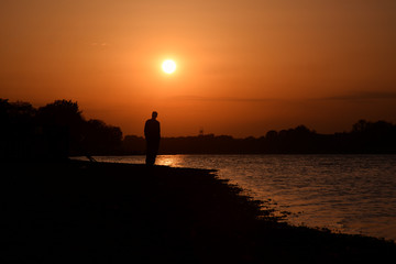 Naklejka na ściany i meble Silhouette of a man in the sunset on the river. The concept of loneliness.