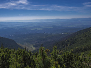 Obraz premium View from tatra mountains trail on valley with Tatranska lomnice and blue misty slopes of hills in the distance. Pine trees and coniferous forest hills, blue sky. Tatra mountain in summer, Slovakia.