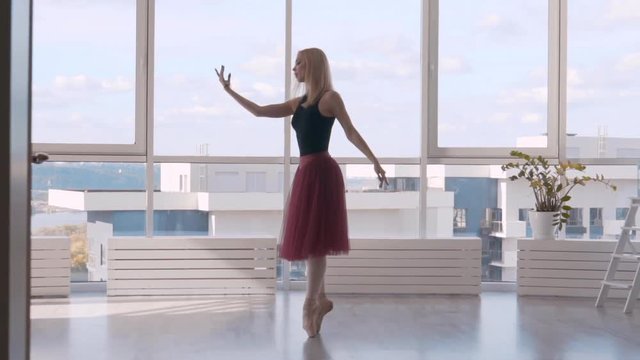 the light and graceful ballerina is dancing in the rays of light against the background of a large window. Woman blonde on pointe in ballet dress. Slowmotion