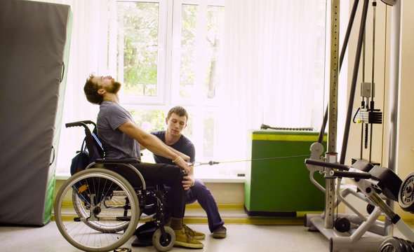 Young disabled man in wheelchair doing strength exercises for hands in the rehabilitation clinic. Doctor physiotherapist helping him
