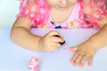 Adorable little girl having fun playing at home with colorful pink nail polish doing manicure and painting nails. Toddler cute child in pink elegant dress sitting wood white table and making manicure