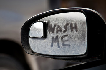 Wash me inscription on the dirty side view mirror of a car