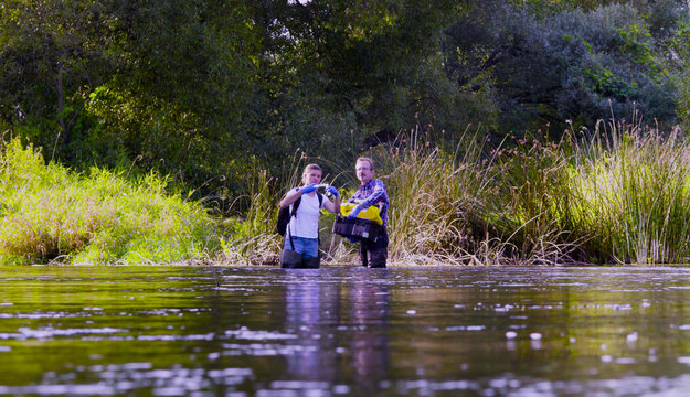 Two scientists ecologists in high rubber boots standing in the water of the forest river. Woman is getting water sampels, man is holding toolbox. Field work