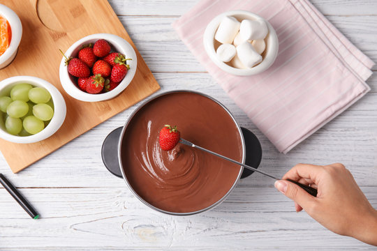 Woman dipping strawberry into pot with chocolate fondue on wooden background, flat lay