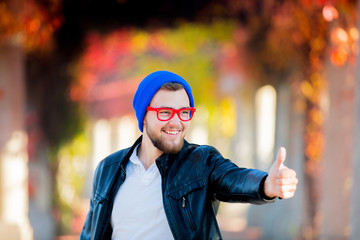 Young white guy in glasses and hat in a park show OK sign with yellow trees on background. Autumn season time