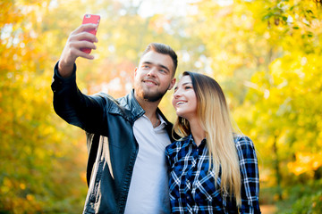 Young white couple make a selfie on mobile phone in a park with yellow trees on background. Autumn season time