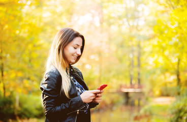 Young white girl using a mobile phone in a park with yellow trees on background. Autumn season time