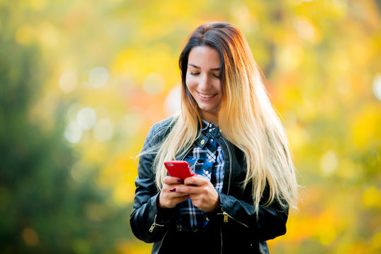 Young white girl using a mobile phone in a park with yellow trees on background. Autumn season time