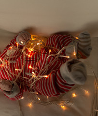 baby in a red striped suit lying on the couch, playing with a garland of included lanterns, Christmas lights, Christmas lights