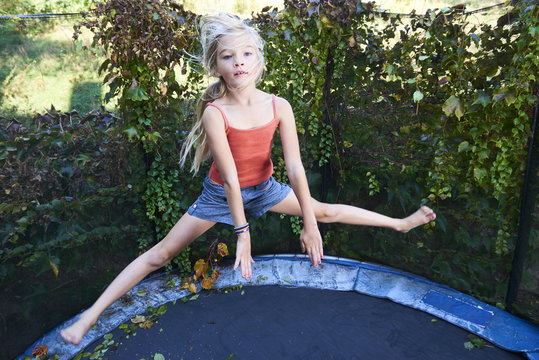 Child cute blond girl playing and jumping on trampoline with greenery background