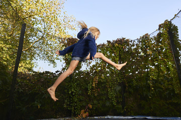 Child cute blond girl playing and jumping on trampoline with greenery background