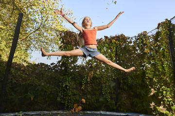 Child cute blond girl playing and jumping on trampoline with greenery background