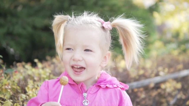 Little girl sucks chupa chups outside in the park. It is autumn, golden leaves. Girl happy with candy in their hands.