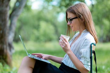 Business woman using laptop on lunch break in city park and hold coffe in her hand