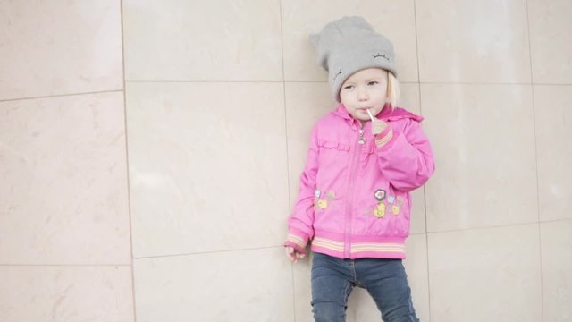 A little girl is standing at the wall of the house and sucking on a candy lollipop. Fashionable video. Baby in a hat and jacket. She really loves this candy.