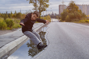 A boy in a dark jacket jumps a puddle on the road