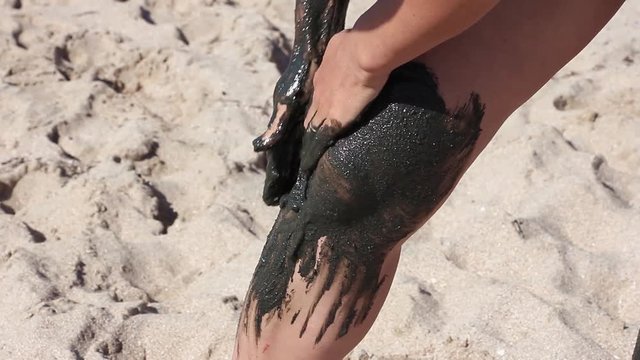 the girl gently covers her leg with therapeutic mud, on the background of sea beach 