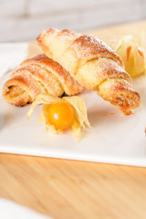 Small croissant with physalis fruits in white ceramic tray, homemade cakes on wooden table top.