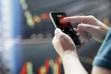 Mans hand holding mobile phone against dark stock market charts