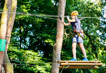 Child in forest adventure park. Kid in orange helmet  and blue t shirt climbs on high rope trail. Agility skills and climbing outdoor amusement center for children. young boy plays outdoors.