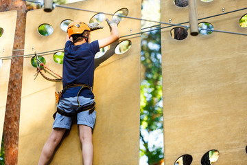 Child in forest adventure park. Kid in orange helmet  and blue t shirt climbs on high rope trail. Agility skills and climbing outdoor amusement center for children. young boy plays outdoors.