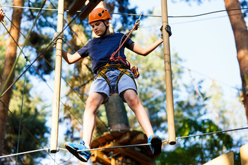 Child in forest adventure park. Kid in orange helmet  and blue t shirt climbs on high rope trail....