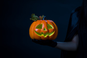 The woman wearing a black dress wearing a black glove hold in orange halloween pumpkin on right hand on the black background