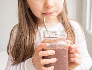 Cropped close up of little girl drinking berry smoothie from glass mug with metal straw (selective...