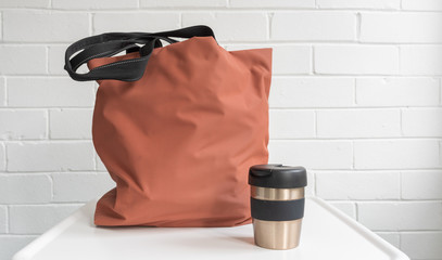 Resuable coffee cup and shopping tote bag on white table against brick wall © Natalie Board