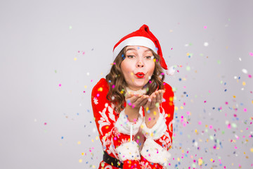 Holiday, Christmas and people concept - Beautiful and young woman in santa claus costume over white background blowing on confetti