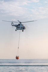 A helicopter with a red basket descends over the sea to scoop water against the background of the dawn orange sky and the silhouette of the city in the distance