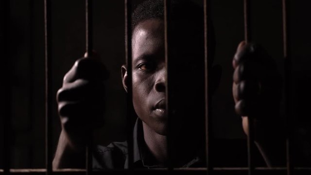 Sad young black american in dark jail,holding bars. Crime,law,punishment