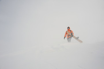 Fototapeta na wymiar Man in the orange sportwear walking with the snowboard in hands in the mountain resort