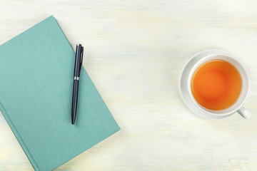 A photo of a teal blue journal with a pen and a cup of tea, shot from above, forming a frame on a light background with copyspace
