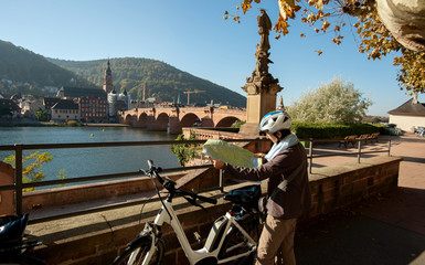 Fototapeta na wymiar Fahrradfahrerin mit Straßenkarte und E-Bike in Heidelberg, mit Blick auf Nepomuk Figur und alte Brücke