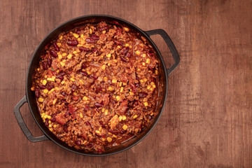 A large cast iron pan with chili con carne, shot from the top on a dark rustic wooden background with copy space