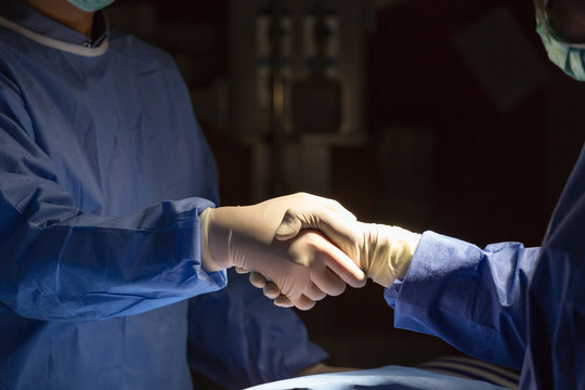 Two surgeons in scrubs shaking hands under surgical light, symbolizing teamwork, trust, and success in medical practice. - Powered by Adobe